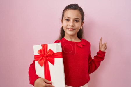 Téléchargez les photos : Petite fille hispanique tenant un cadeau souriant heureux pointant avec la main et le doigt sur le côté - en image libre de droit