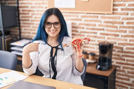 Téléchargez les photos : Jeune fille aux cheveux bleus tenant modèle d'organe génital féminin au bureau se plaignant de douleur menstruelle pointant du doigt un sourire heureux et fier - en image libre de droit