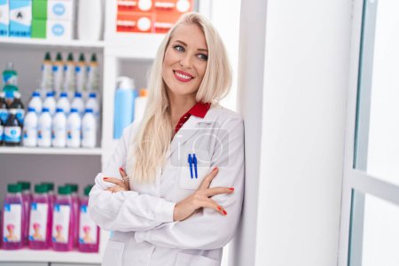 Foto de Joven mujer rubia farmacéutica sonriendo confiada de pie con los brazos cruzados gesto en la farmacia - Imagen libre de derechos