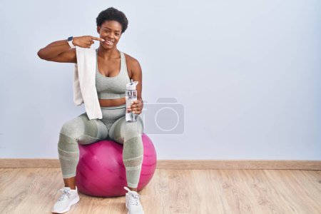 Téléchargez les photos : Femme afro-américaine portant des vêtements de sport assis sur pilates balle souriant gai montrant et pointant avec les doigts dents et la bouche. concept de santé dentaire. - en image libre de droit