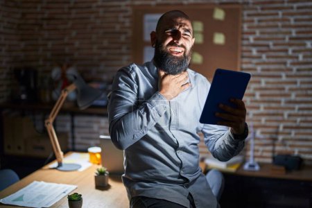 Téléchargez les photos : Jeune homme hispanique avec barbe et tatouages travaillant au bureau la nuit touchant le cou douloureux, mal de gorge pour la grippe, la motte et l'infection - en image libre de droit