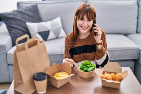 Photo for Young woman talking on the smartphone eating take away food at home - Royalty Free Image