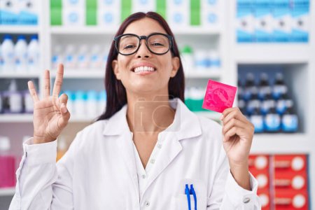 Téléchargez les photos : Jeune femme hispanique travaillant à la pharmacie tenant préservatif faire ok signe avec les doigts, sourire geste amical excellent symbole - en image libre de droit