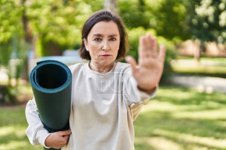 Foto de Mujer hispana de mediana edad sosteniendo esterilla de yoga en el parque con la mano abierta haciendo stop sign con expresión seria y confiada, gesto de defensa - Imagen libre de derechos
