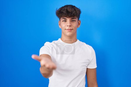 Téléchargez les photos : Adolescent hispanique debout sur fond bleu souriant joyeuse offrant main de paume donnant assistance et acceptation. - en image libre de droit