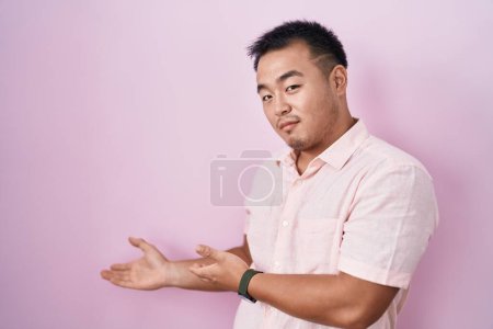 Foto de Chino joven de pie sobre fondo rosa invitando a entrar sonriente natural con la mano abierta - Imagen libre de derechos