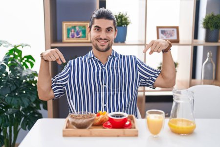 Foto de Hombre hispano con el pelo largo sentado en la mesa desayunando confiado con sonrisa en la cara, señalándose con los dedos orgullosos y felices. - Imagen libre de derechos