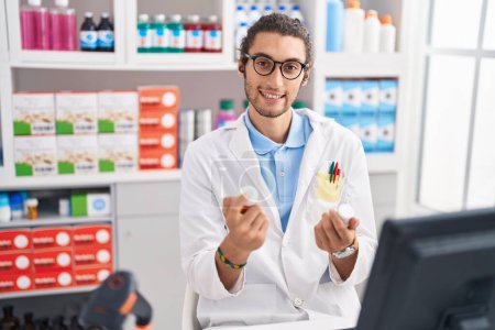 Téléchargez les photos : Young hispanic man pharmacist smiling confident holding pills bottles at pharmacy - en image libre de droit