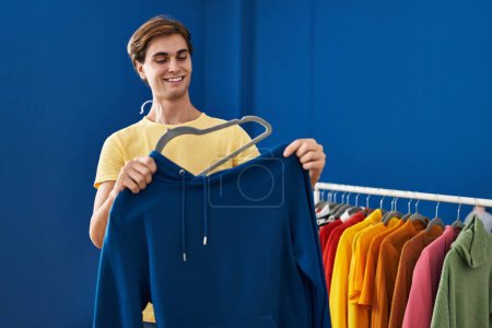 Téléchargez les photos : Jeune homme caucasien souriant confiant mettre sweat-shirt sur cintre à la buanderie - en image libre de droit