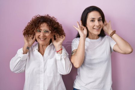 Foto de Madre e hija hispanas vistiendo una camiseta blanca casual sobre fondo rosa tratando de escuchar el gesto de ambas manos en la oreja, curiosas de chismes. problema auditivo, sordo - Imagen libre de derechos
