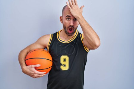 Téléchargez les photos : Jeune homme chauve avec barbe portant l'uniforme de basket-ball tenant ballon surpris avec la main sur la tête pour erreur, rappelez-vous erreur. oublié, mauvais concept de mémoire. - en image libre de droit
