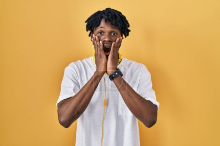 Téléchargez les photos : Jeune homme africain avec dreadlocks debout sur fond jaune effrayé et choqué, expression surprise et stupéfaite avec les mains sur le visage - en image libre de droit