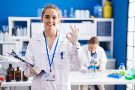 Foto de Mujer rubia que trabaja en el laboratorio científico haciendo signo ok con los dedos, sonriendo gesto amistoso excelente símbolo - Imagen libre de derechos