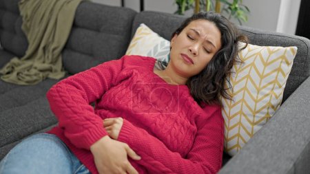 Foto de Joven mujer hispana hermosa que sufre de dolor de estómago tumbada en el sofá en casa - Imagen libre de derechos