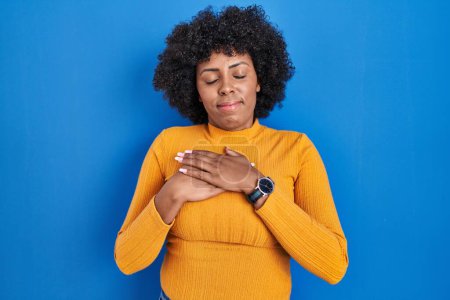 Photo for Black woman with curly hair standing over blue background smiling with hands on chest with closed eyes and grateful gesture on face. health concept. - Royalty Free Image