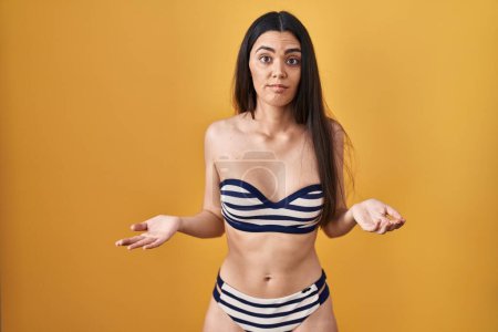Foto de Mujer morena joven con bikini sobre fondo amarillo despistado y confundido con los brazos abiertos, sin concepto de idea. - Imagen libre de derechos