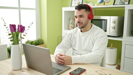 Photo for Young hispanic man using laptop and headphones sitting on table at dinning room - Royalty Free Image