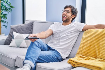 Foto de Hombre latino guapo sosteniendo el control remoto de la televisión sonriendo y riendo en voz alta porque divertido chiste loco. - Imagen libre de derechos