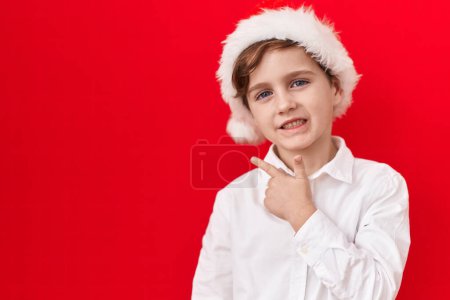 Photo for Little caucasian boy wearing christmas hat over red background smiling cheerful pointing with hand and finger up to the side - Royalty Free Image