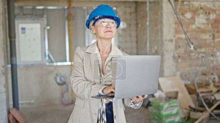 Foto de Arquitecta rubia de mediana edad usando laptop con expresión relajada en el sitio de construcción - Imagen libre de derechos
