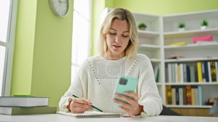 Téléchargez les photos : Jeune étudiante blonde utilisant un smartphone prenant des notes à la bibliothèque de l'université - en image libre de droit