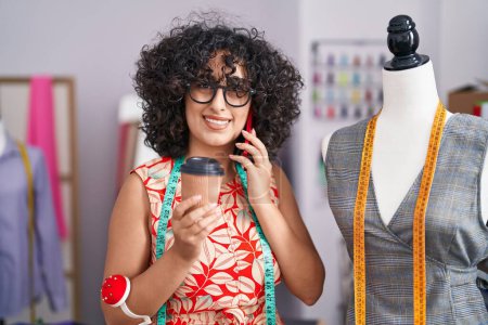 Téléchargez les photos : Jeune femme du Moyen-Orient tailleur parler sur smartphone boire du café au magasin de tailleur - en image libre de droit