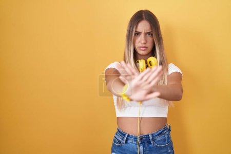 Foto de Mujer rubia joven de pie sobre fondo amarillo con auriculares expresión de rechazo cruzando brazos y palmas haciendo signo negativo, cara enojada - Imagen libre de derechos