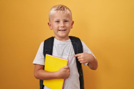 Téléchargez les photos : Petit garçon caucasien portant sac à dos étudiant et tenant livre souriant heureux pointant avec la main et le doigt - en image libre de droit