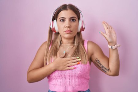 Photo for Young blonde woman listening to music using headphones swearing with hand on chest and open palm, making a loyalty promise oath - Royalty Free Image
