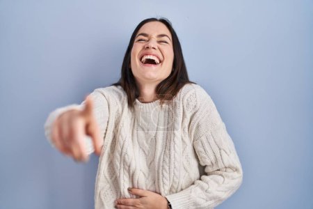 Photo for Young brunette woman standing over blue background laughing at you, pointing finger to the camera with hand over body, shame expression - Royalty Free Image