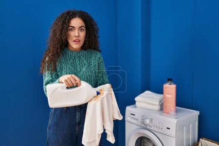 Foto de Mujer hispana joven sosteniendo ropa sucia y botella de detergente expresión despistada y confusa. concepto de duda. - Imagen libre de derechos