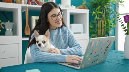 Téléchargez les photos : Jeune femme hispanique avec chihuahua chien en utilisant un ordinateur portable à la salle à manger - en image libre de droit