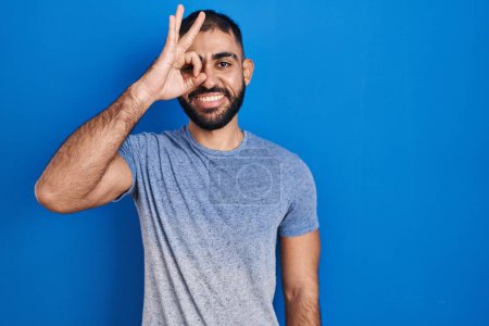 Téléchargez les photos : Middle east man with beard standing over blue background doing ok gesture with hand smiling, eye looking through fingers with happy face. - en image libre de droit