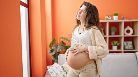 Téléchargez les photos : Jeune femme enceinte massant le ventre debout à la maison - en image libre de droit