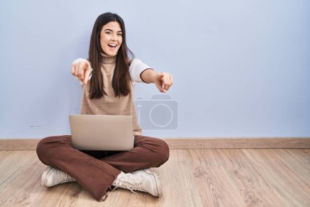 Foto de Mujer morena joven trabajando con computadora portátil sentada en el suelo señalándote a ti y a la cámara con los dedos, sonriendo positiva y alegre - Imagen libre de derechos