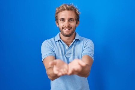 Téléchargez les photos : Homme caucasien debout sur fond bleu souriant avec les mains paumes ensemble recevant ou donnant geste. maintien et protection - en image libre de droit