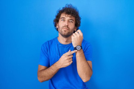 Téléchargez les photos : Jeune homme hispanique debout sur fond bleu pressé pointant vers regarder le temps, l'impatience, regardant la caméra avec une expression détendue - en image libre de droit