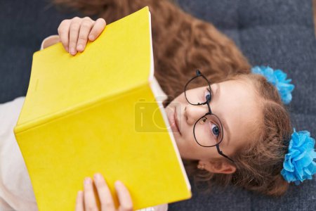 Foto de Adorable chica caucásica leyendo libro acostado en el sofá en casa - Imagen libre de derechos
