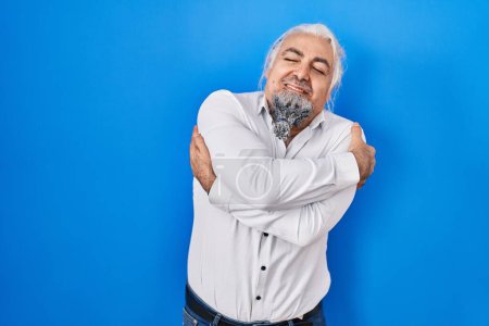 Téléchargez les photos : Homme d'âge moyen avec les cheveux gris debout sur fond bleu s'étreignant heureux et positif, souriant confiant. amour de soi et soins de soi - en image libre de droit