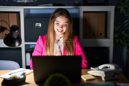 Téléchargez les photos : Young caucasian woman working at the office at night praying with hands together asking for forgiveness smiling confident. - en image libre de droit