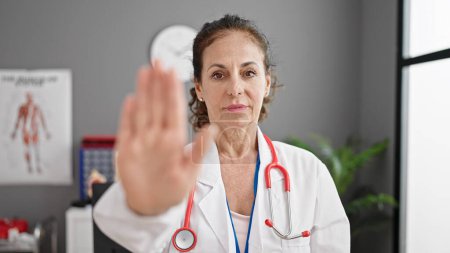 Téléchargez les photos : Moyen âge hispanique femme médecin faire stop signe avec la main à la clinique - en image libre de droit