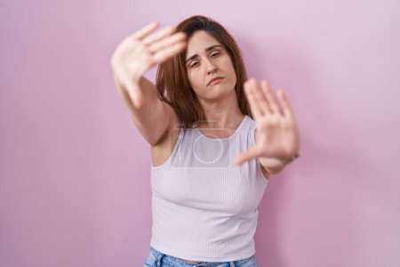 Photo for Brunette woman standing over pink background doing frame using hands palms and fingers, camera perspective - Royalty Free Image