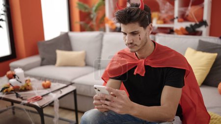 Foto de Joven hombre hispano usando disfraz de diablo usando smartphone en casa - Imagen libre de derechos