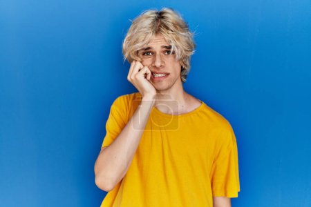 Foto de Joven hombre moderno de pie sobre fondo azul mirando estresado y nervioso con las manos en la boca mordiendo las uñas. problema de ansiedad. - Imagen libre de derechos