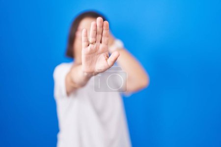 Foto de Mujer hispana de mediana edad de pie sobre fondo azul cubriendo los ojos con las manos y haciendo un gesto de stop con expresión triste y de miedo. concepto avergonzado y negativo. - Imagen libre de derechos