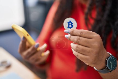 Photo for African american woman using smartphone holding bitcoin at office - Royalty Free Image