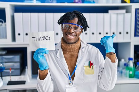 Foto de Joven africano con rastas trabajando en laboratorio científico apuntando con el pulgar hacia un lado sonriendo feliz con la boca abierta - Imagen libre de derechos