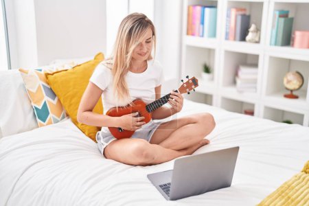Téléchargez les photos : Jeune femme blonde ayant une leçon d'ukulélé en ligne assis sur le lit dans la chambre - en image libre de droit