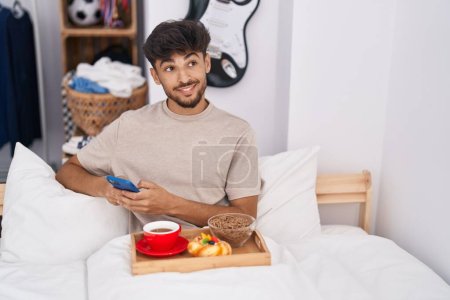 Foto de Hombre árabe joven usando teléfono inteligente desayunando en el dormitorio - Imagen libre de derechos