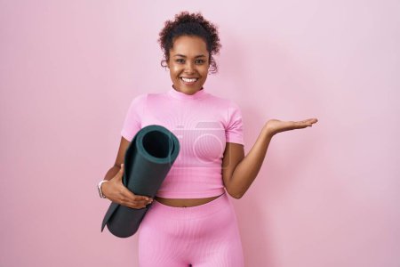 Foto de Joven mujer hispana con pelo rizado sosteniendo esterilla de yoga sobre fondo rosa sonriente alegre presentando y señalando con la palma de la mano mirando a la cámara. - Imagen libre de derechos